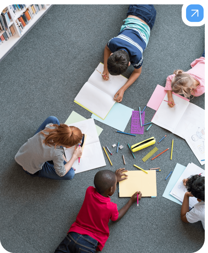 Mehrere Kinder liegen auf dem Teppichboden einer Bibliothek oder eines Klassenraums und malen oder schreiben in ihre Hefte. Um sie herum liegen bunte Stifte, Lineale, Papier und eine Federmappe verstreut. Die Atmosphäre wirkt kreativ und konzentriert, die Kinder sind in ihre Aufgaben vertieft. Im Hintergrund sind Bücherregale zu sehen.