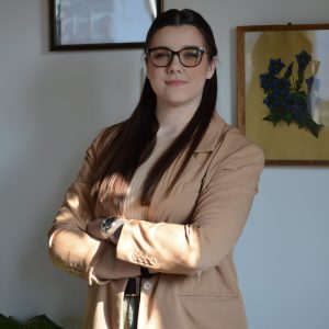 Eine Person mit langen, braunen Haaren steht mit verschränkten Armen in einem Raum. Sie trägt ein beigefarbenes Sakko über einem passenden Oberteil sowie eine Armbanduhr. Im Hintergrund hängen zwei Bilder an der Wand, eines davon zeigt blaue Blumen. Die Atmosphäre wirkt ruhig und professionell.
