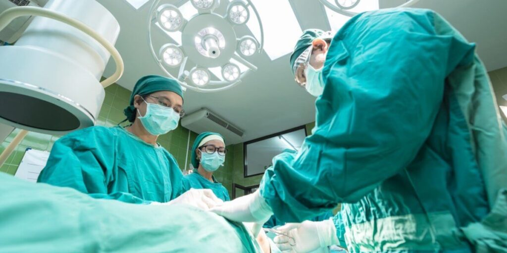 Mehrere Personen in grüner OP-Kleidung, einschließlich Hauben, Masken und Handschuhen, führen gemeinsam einen chirurgischen Eingriff in einem Operationssaal durch. Über dem Operationstisch hängt eine große, helle OP-Lampe. Im Hintergrund sind medizinische Geräte und grün gekachelte Wände zu sehen.
