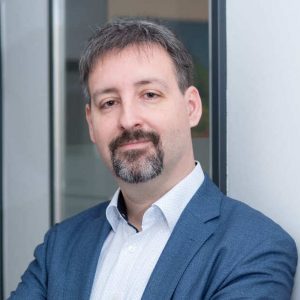 Eine Person steht mit verschränkten Armen in einem modernen Bürogebäude. Sie trägt ein blaues Sakko über einem weißen Hemd. Im Hintergrund sind Glaswände und eine helle Wand zu sehen. Die Haltung wirkt professionell und selbstbewusst.