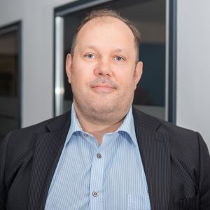 Eine Person steht in einem modernen Büroraum. Sie trägt ein hellblaues, gestreiftes Hemd und ein dunkel gestreiftes Sakko. Im Hintergrund sind Fenster mit dunklen Rahmen und helle Wände zu sehen. Die Atmosphäre wirkt professionell und geschäftlich.