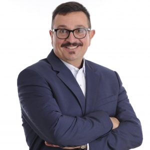 Eine Person steht vor einem weißen Hintergrund und trägt einen dunkelblauen Anzug mit einem weißen Hemd. Die Arme sind verschränkt. Das Auftreten wirkt professionell und selbstbewusst, wie bei einem klassischen Business-Porträt.