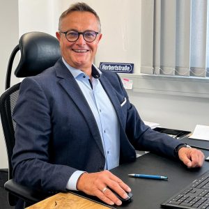 Eine Person sitzt an einem Schreibtisch in einem modernen Büro. Sie trägt ein dunkelblaues Sakko über einem hellblauen Hemd und hat einen Ring am linken Ringfinger. Die rechte Hand hält eine Computermaus, vor ihr liegt eine Tastatur. Auf dem Schreibtisch befinden sich außerdem ein Kugelschreiber, einige Unterlagen und ein kleines Straßenschild mit der Aufschrift „Herbertstraße“. Im Hintergrund sind graue Jalousien am Fenster zu sehen. Die Atmosphäre wirkt professionell und strukturiert.