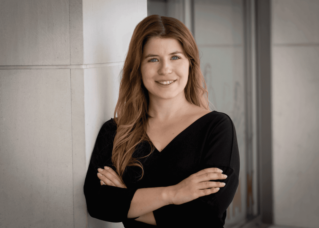 Eine Person mit langen, leicht gewellten, braunen Haaren steht mit verschränkten Armen an eine helle Wand gelehnt. Sie trägt ein schwarzes Oberteil mit V-Ausschnitt. Im Hintergrund ist ein moderner, heller Innenraum zu erkennen. Die Haltung wirkt selbstbewusst und professionell.