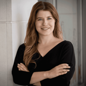 Eine Person mit langen, leicht gewellten, braunen Haaren steht mit verschränkten Armen an eine helle Wand gelehnt. Sie trägt ein schwarzes Oberteil mit V-Ausschnitt. Im Hintergrund ist ein moderner, heller Innenraum zu erkennen. Die Haltung wirkt selbstbewusst und professionell.