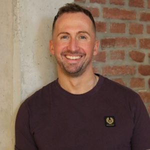 Eine Person steht vor einer Wand, die aus unverputzten Ziegelsteinen und einem Teil aus hellem, rauem Putz besteht. Sie trägt ein dunkelviolettes T-Shirt mit einem kleinen, rechteckigen Patch der Marke Belstaff auf der Brust. Die Atmosphäre wirkt lässig und urban.