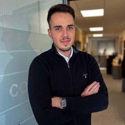 Eine Person steht mit verschränkten Armen in einem modernen Büroflur. Sie trägt einen schwarzen Pullover mit Reißverschluss und ein weißes Hemd darunter. Am linken Handgelenk befindet sich eine Armbanduhr. Im Hintergrund sind Büros und Arbeitsplätze zu erkennen. Auf der Glaswand neben der Person ist der Schriftzug „COVAGO“ teilweise sichtbar. Die Atmosphäre wirkt professionell und freundlich.